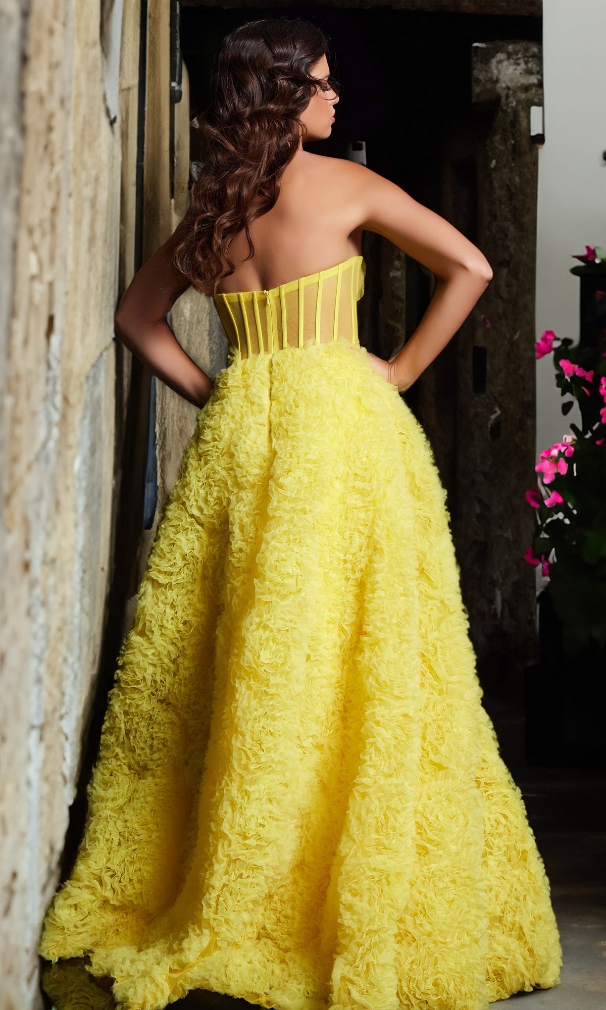 A woman stands with her back to the camera, hands on hips, wearing the Jovani Strapless Long Rosette Prom Dress 38674 with a bright yellow textured skirt, in a stone-walled hallway near pink flowers.